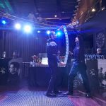 Two barman doing a trick with drinks while holding multiple glasses in their hands.